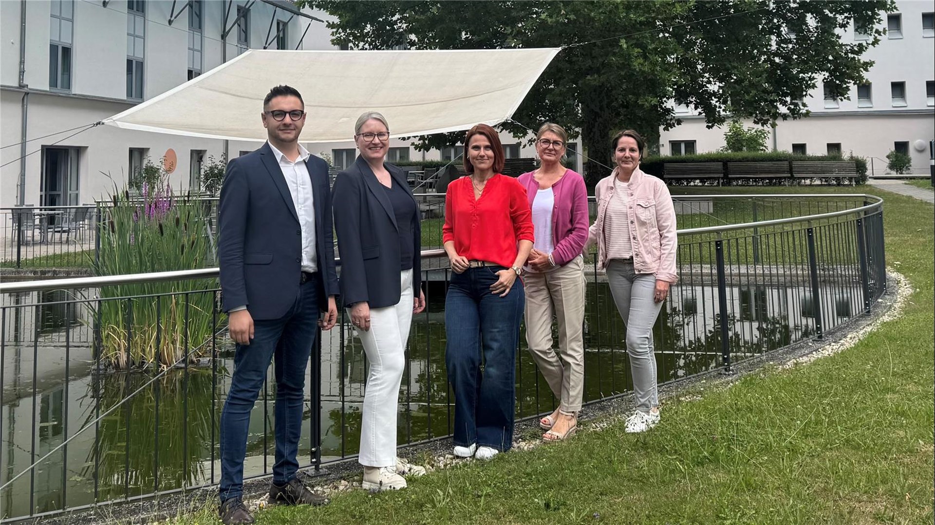 Fünf Personen stehen nebeneinander vor einem kleinen Teich mit Geländer im Außenbereich eines modernen Gebäudes. Sie lächeln in die Kamera.
