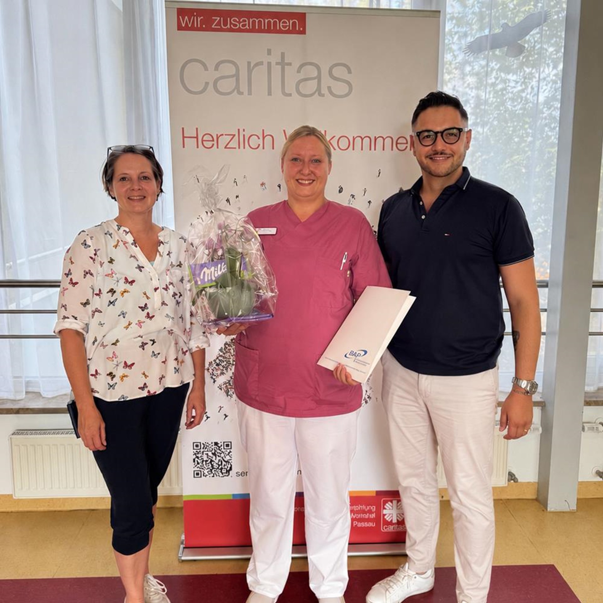 Drei Personen stehen vor einem Caritas Logo. Links zwei Frauen und rechst ein Mann.